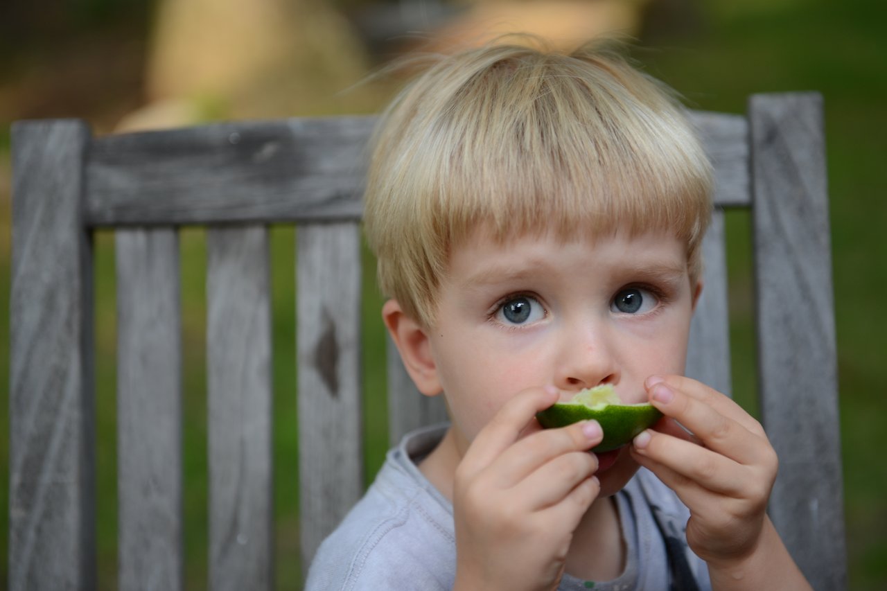 A young child sits on a wooden chair, biting into a green fruit while looking ahead.