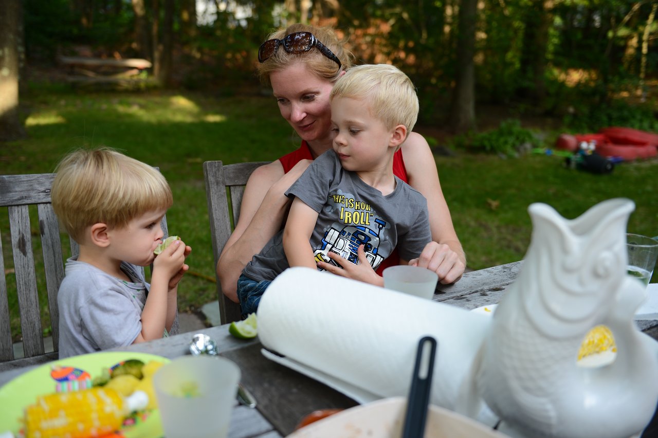 A woman sits at a picnic table holding a young boy, while another child eats nearby.