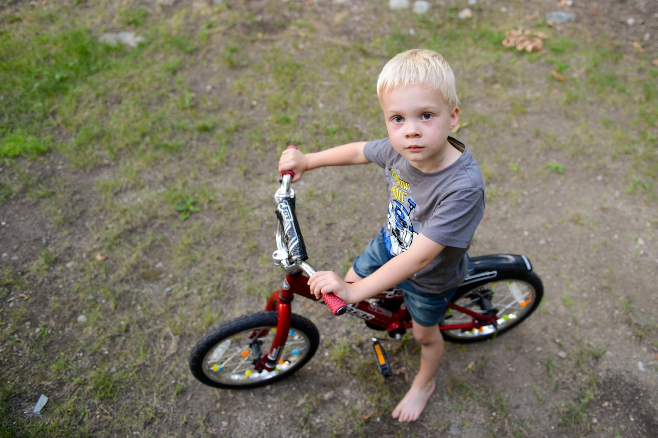 A young child stands barefoot on a red bicycle, looking up while holding the handlebars in a grassy backyard.