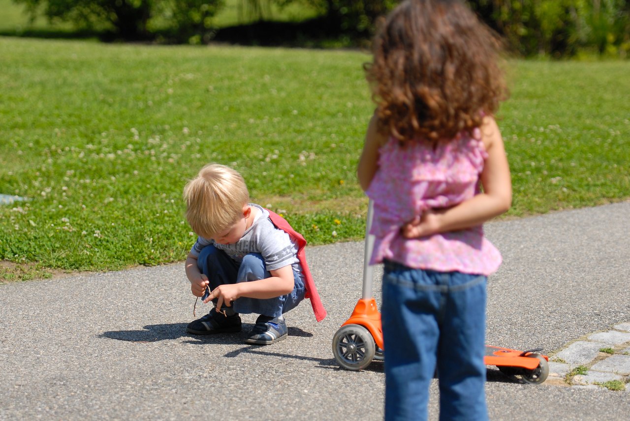 A girl stands near her scooter, watching a boy crouched on the ground examining something in his hands.
