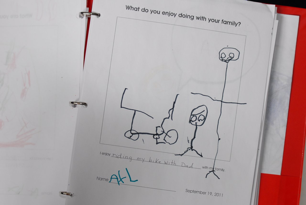 A child's drawing in a binder shows a family activity with a caption about riding a bike with Dad.