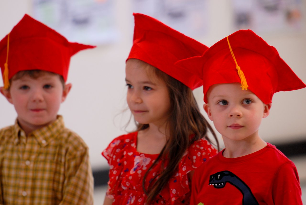 Three young children wearing red graduation caps stand together, looking forward with neutral expressions.