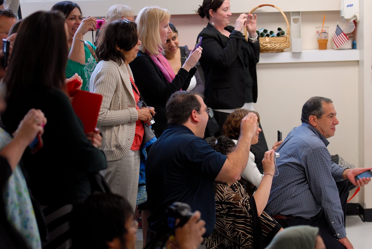 A group of people stand and kneel, holding phones and cameras, capturing a moment at a graduation event.