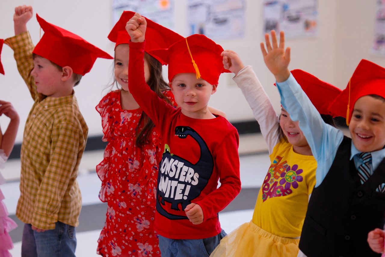A group of young children wearing red graduation caps raise their hands and smile during a celebration.