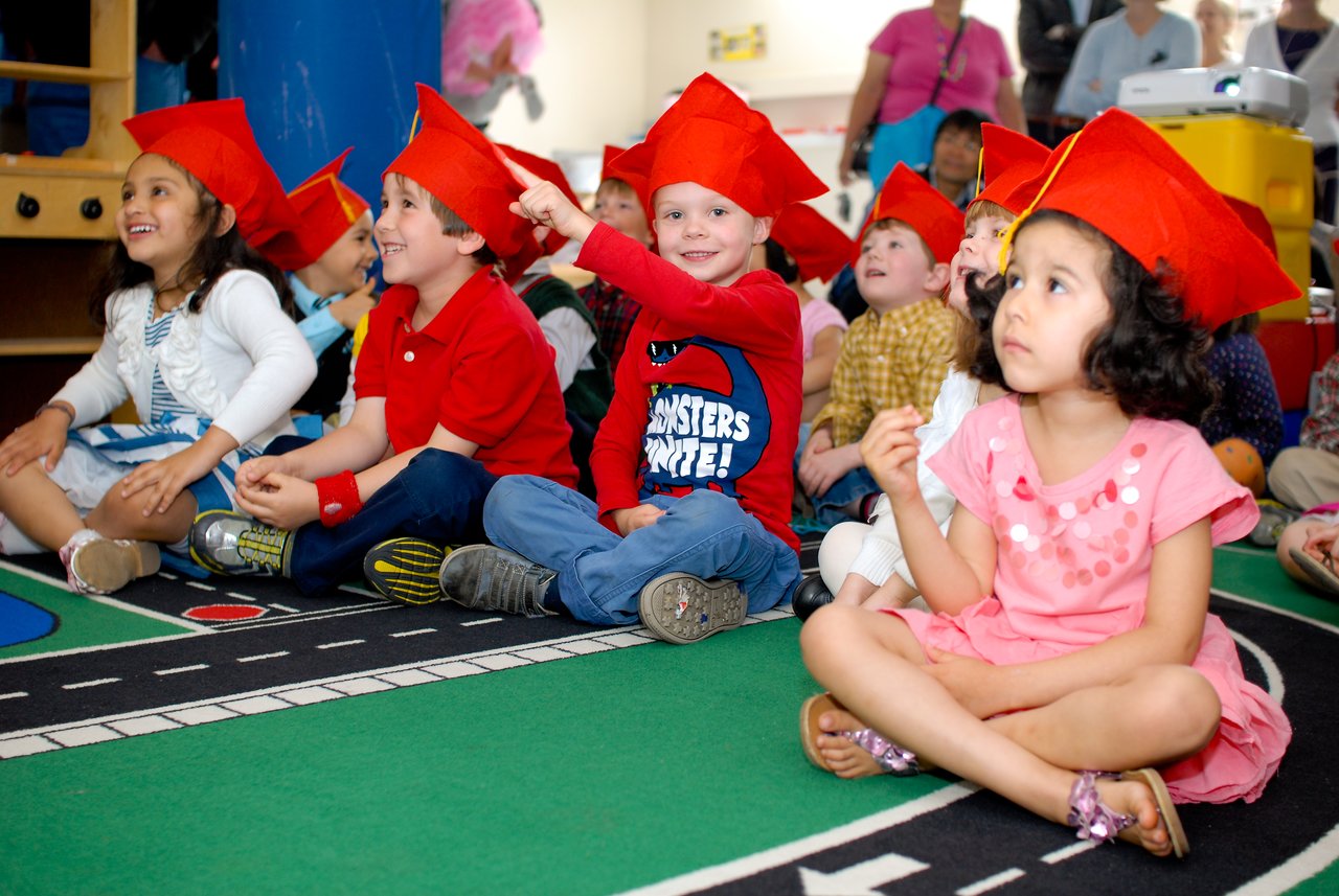 A group of young children wearing red graduation caps sit on the floor, smiling and raising their hands.