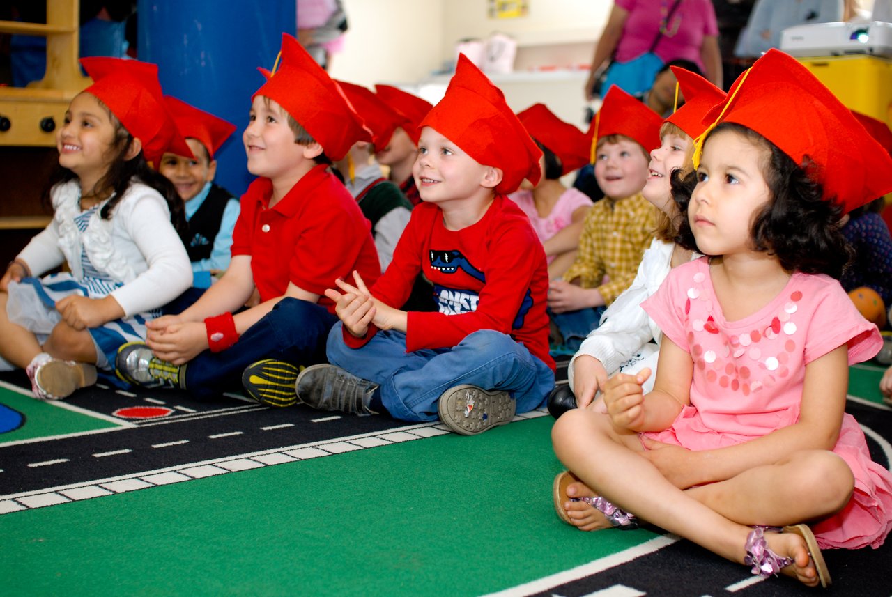 A group of young children wearing red graduation caps sit on the floor, smiling and watching something off-camera.