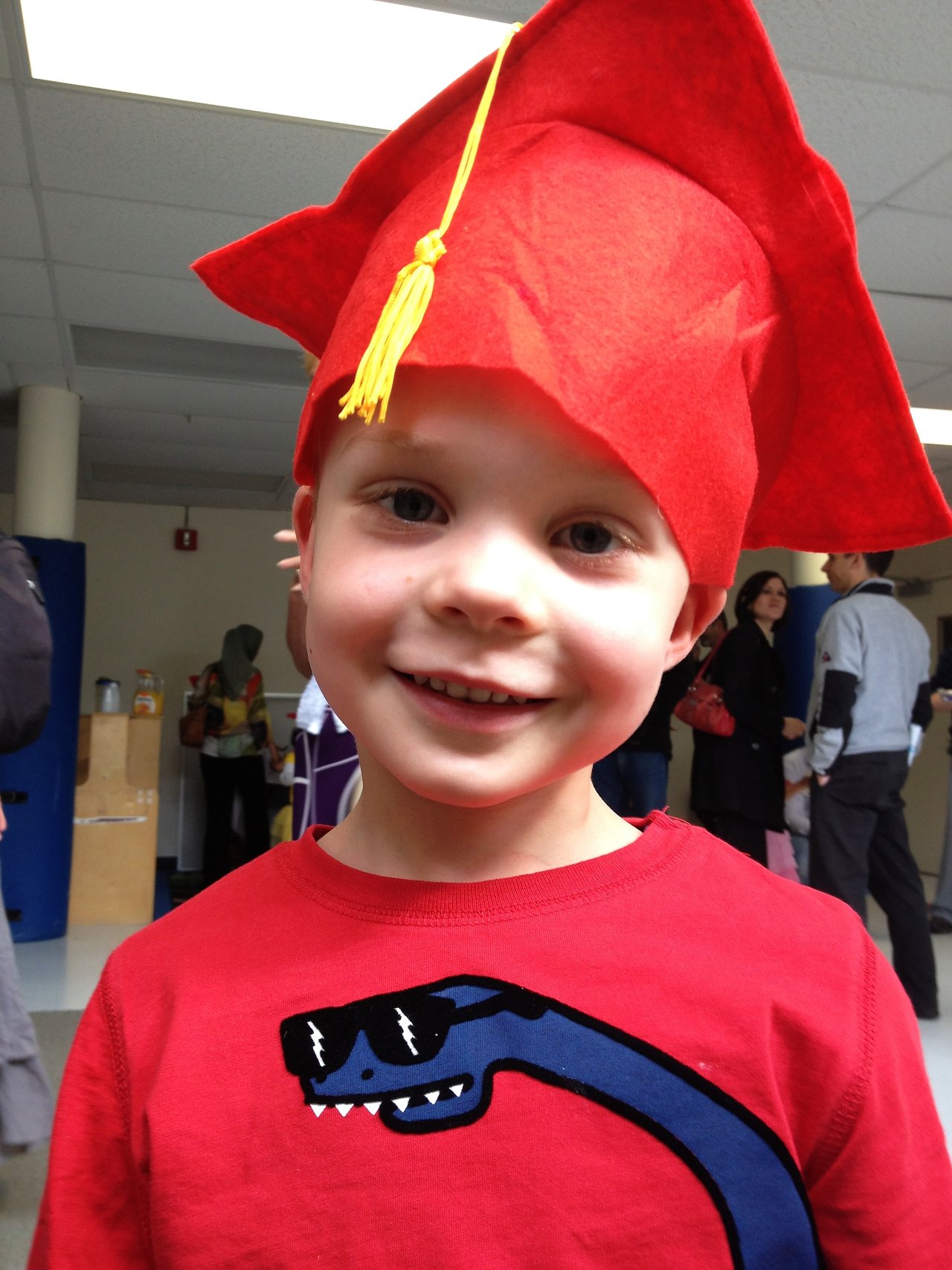 A young child wearing a red graduation cap smiles at the camera in a busy indoor setting.
