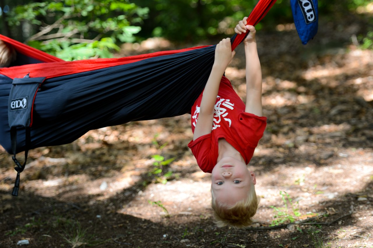 A child in a red shirt hangs upside down from a hammock, holding onto the fabric with both hands.