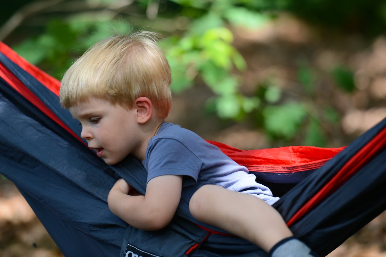 A young child in a blue shirt climbs onto a red and black hammock outdoors, gripping the fabric for balance.
