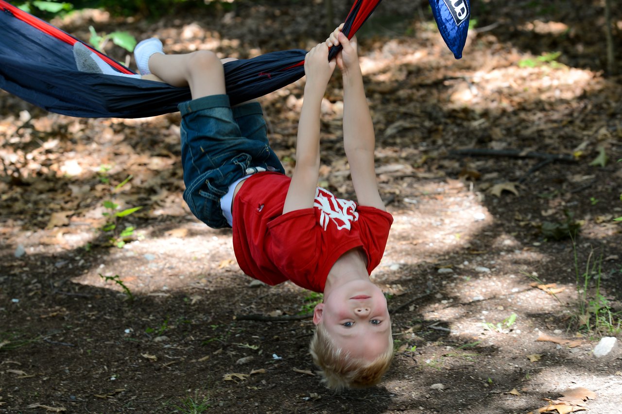 A child in a red shirt hangs upside down from a hammock, gripping the fabric with both hands.