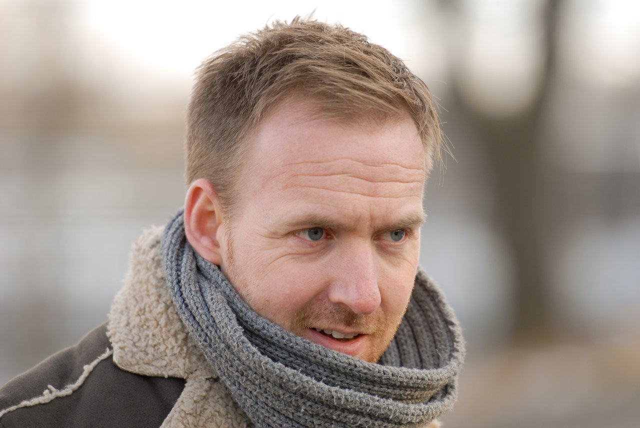 A man wearing a warm coat and scarf looks slightly to the side with a focused expression.