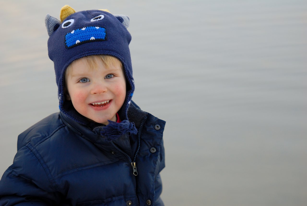 A smiling child in a blue jacket and monster hat stands near a calm body of water.