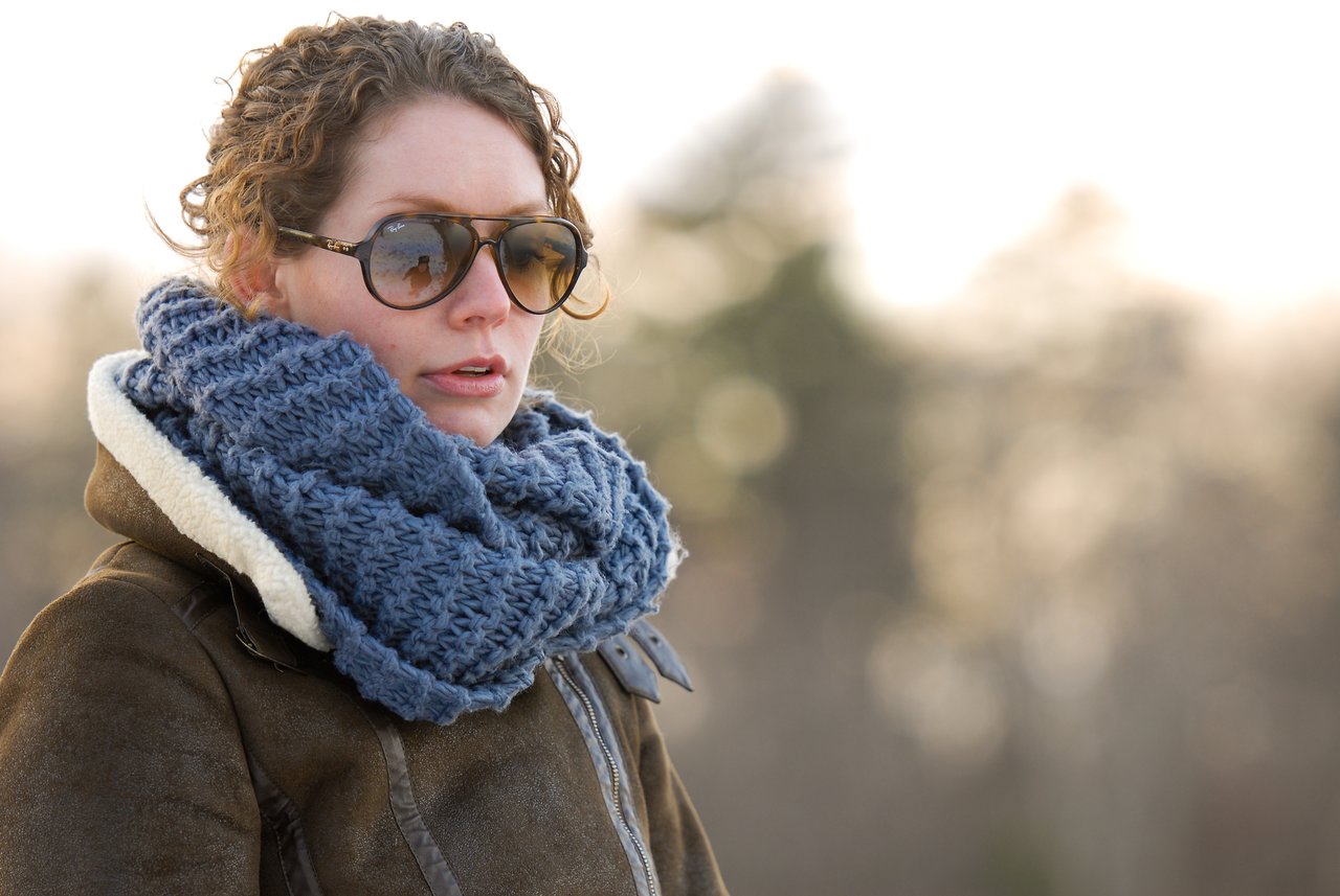 A person wearing sunglasses, a thick blue scarf, and a brown coat stands outdoors in cool weather.