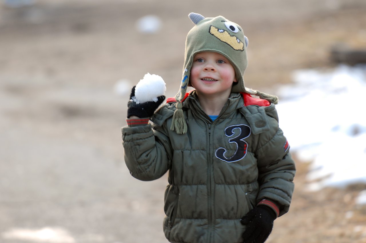A young child in a winter coat and hat holds a snowball while smiling outdoors.