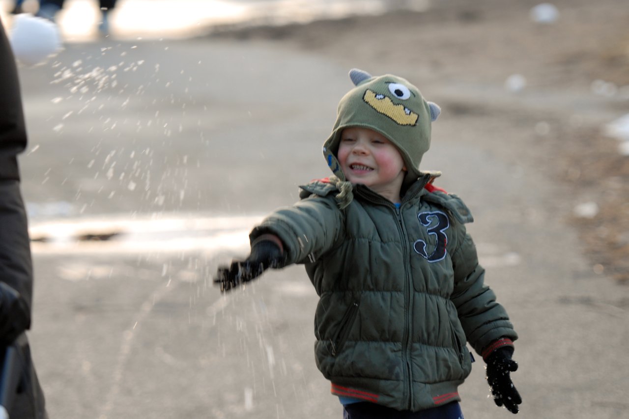 A young child in a green jacket and monster hat throws snow while smiling on a winter day.