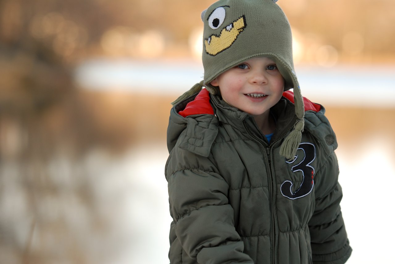 A young child in a green winter coat and playful hat smiles while outdoors on a cold day.