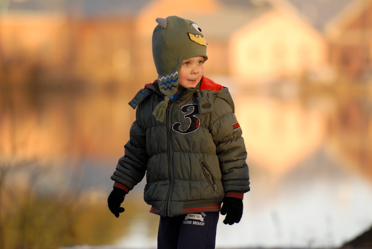 A young child in a winter coat and hat stands outdoors near a pond, looking to the side.