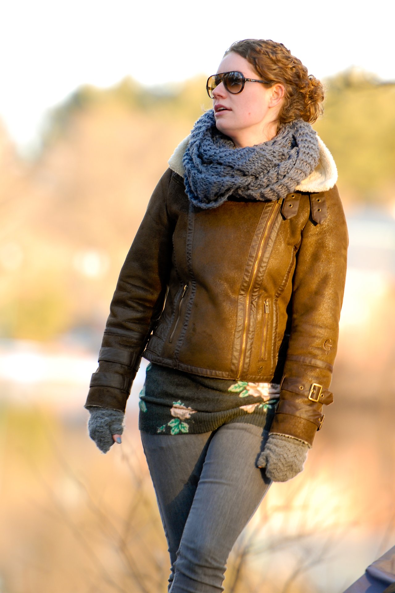 A woman wearing a brown jacket, scarf, and gloves walks outdoors on a cool day, looking to the side.