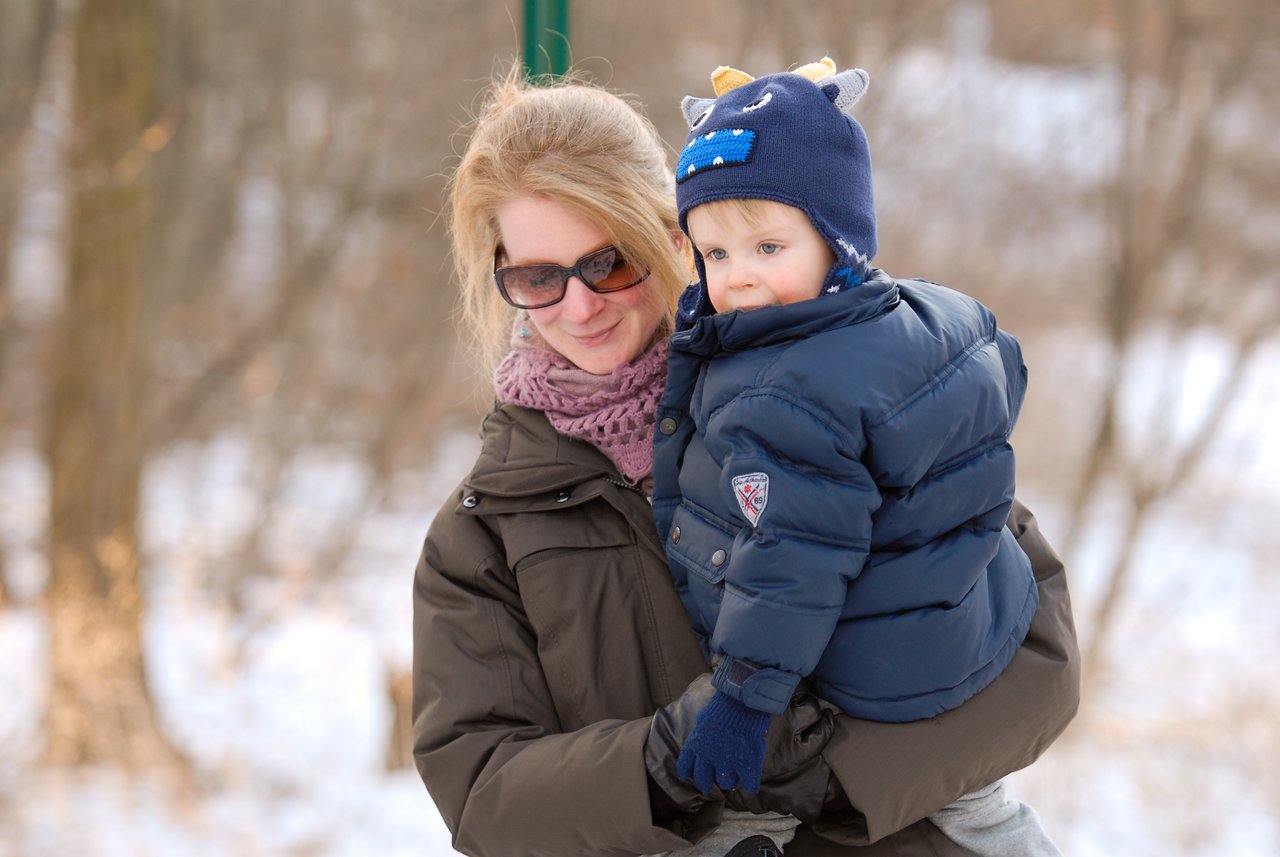 A woman wearing sunglasses holds a young child dressed in a puffy jacket and a hat with ears outdoors.
