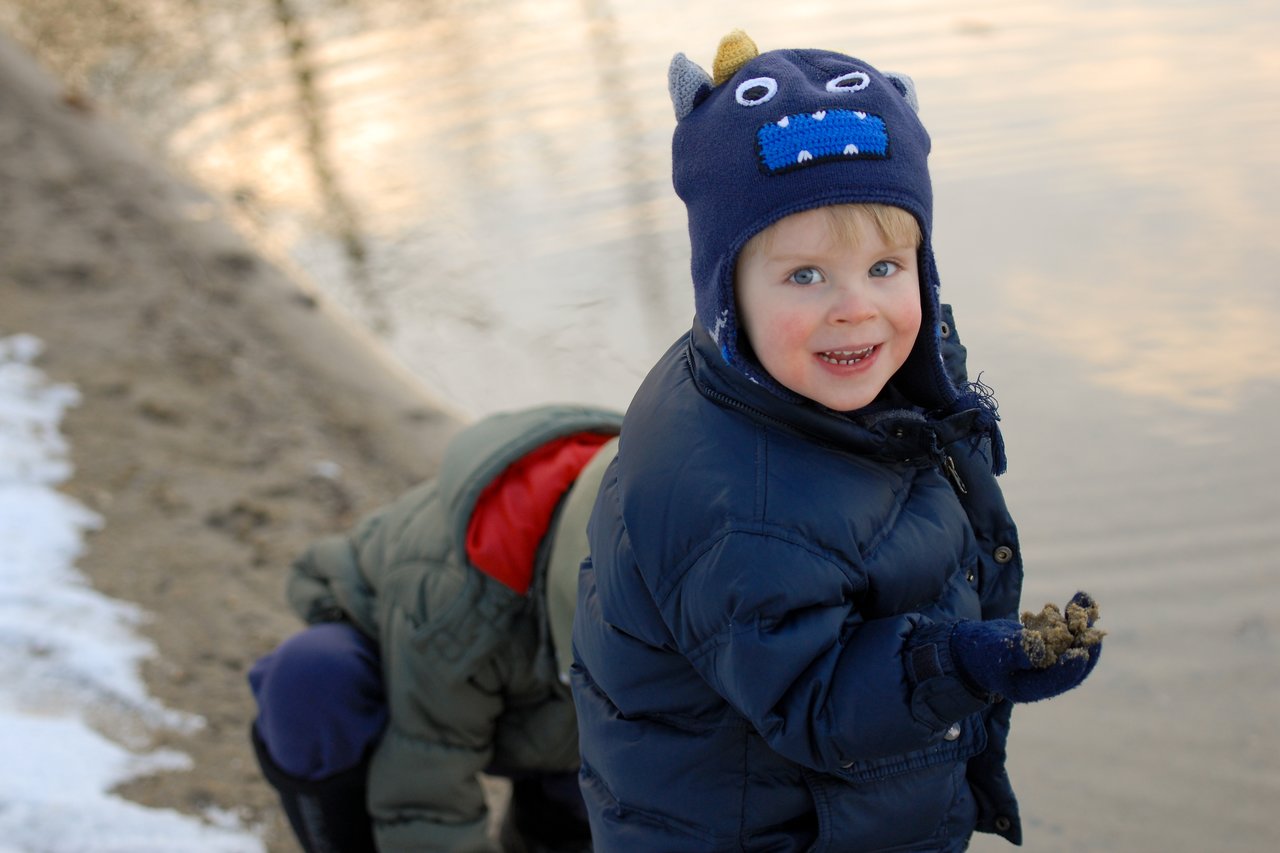 A child in a blue jacket and monster hat smiles while holding sand near the water, another child bends nearby.