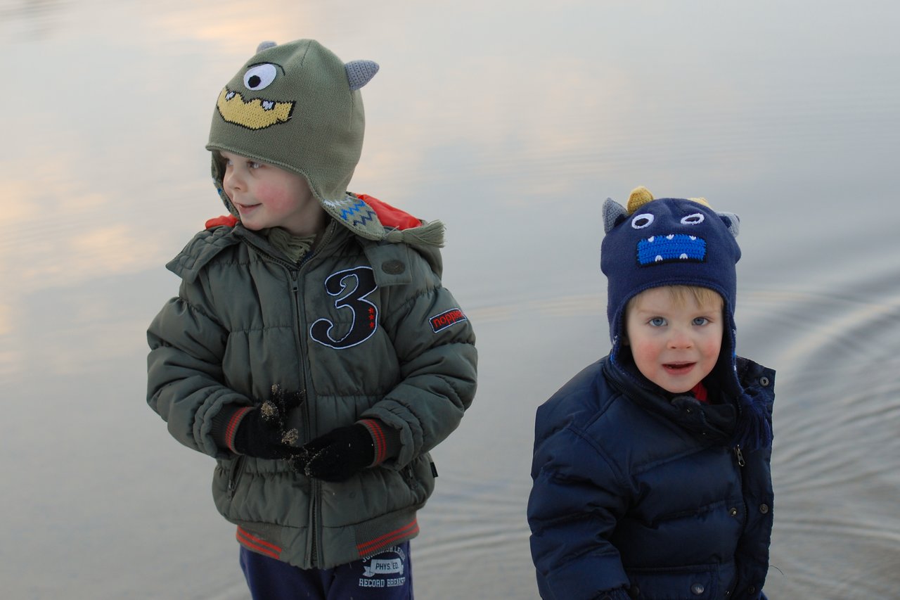 Two young children wearing monster-themed hats stand near the water, dressed in warm jackets and gloves.