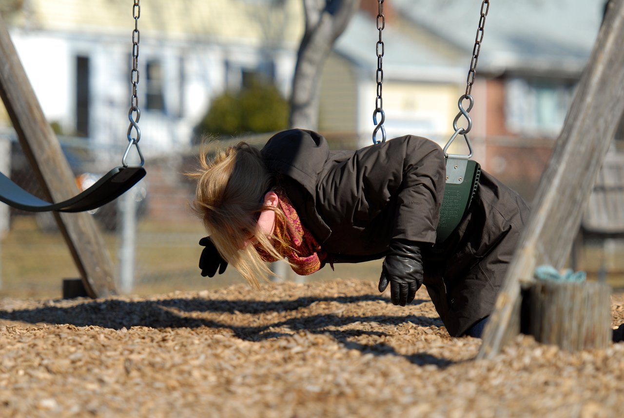 A person in a winter coat leans forward on a swing, arms outstretched, appearing to imitate flying.