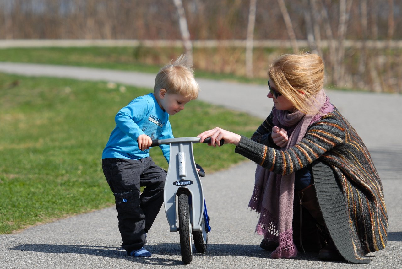 A woman helps a young child balance on a small bike while kneeling on a paved path.