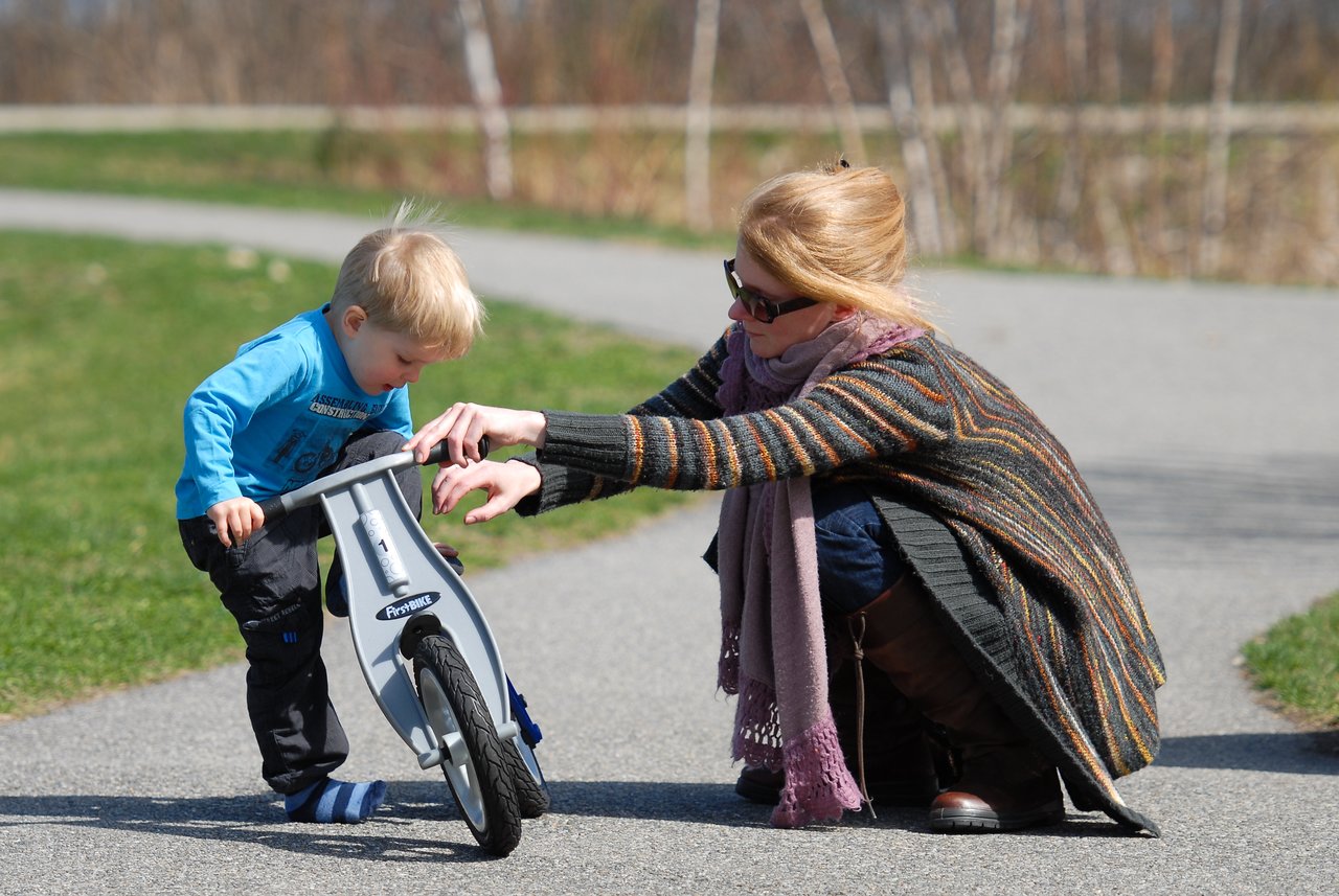 A woman helps a young child balance on a small bike while crouching on a paved path outdoors.