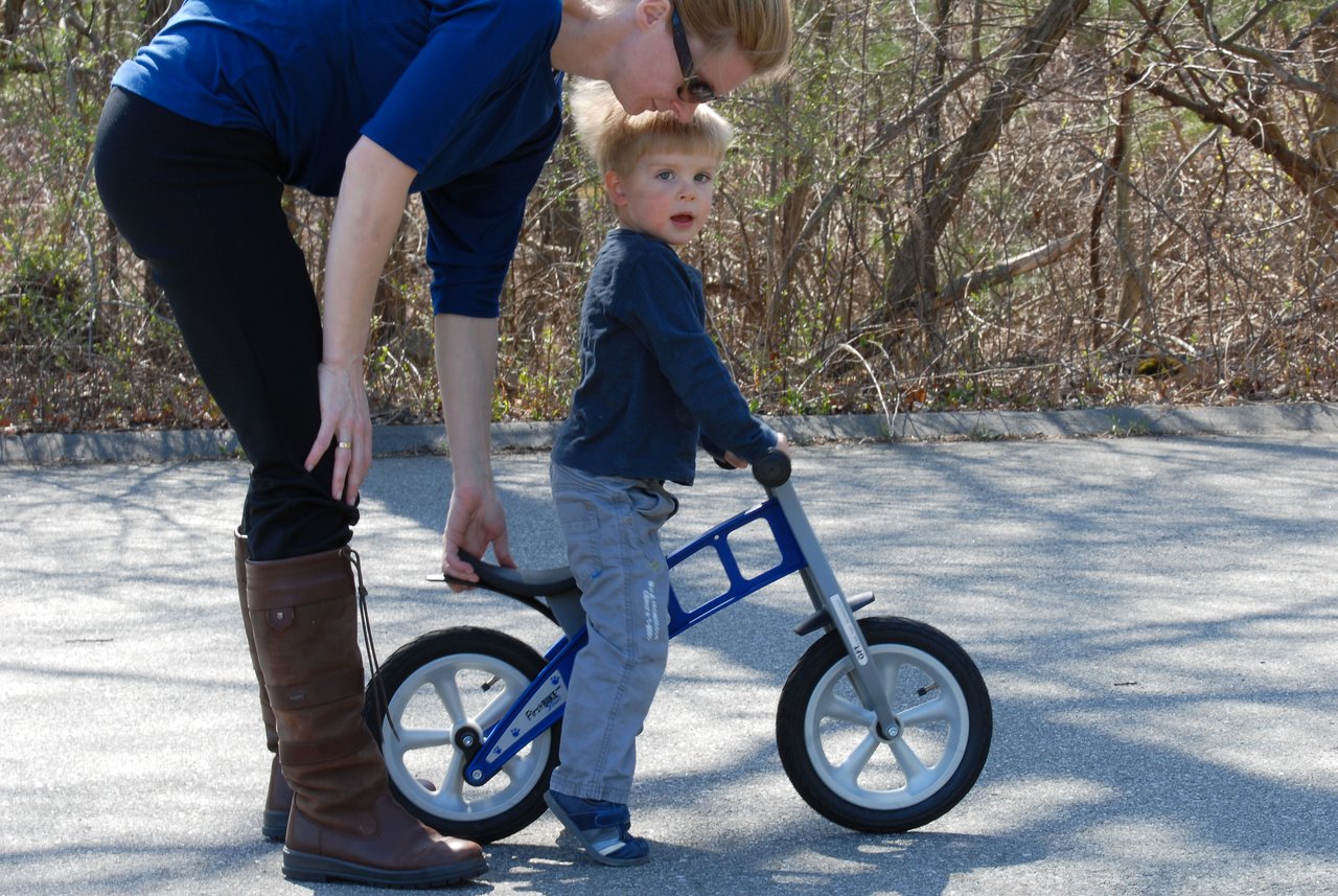 A woman helps a young child balance on a small blue bike while standing on a paved path.