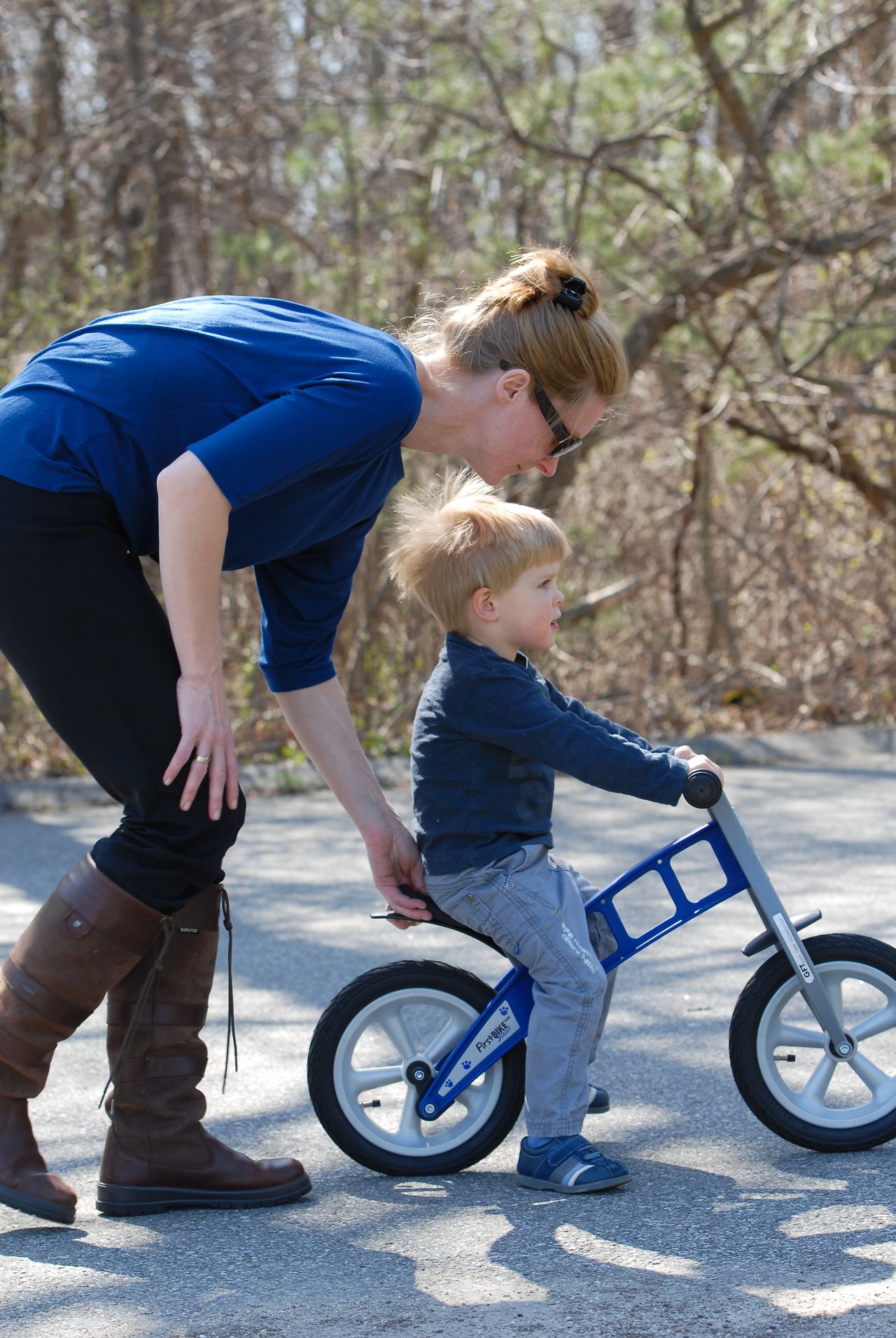 A woman helps a young child balance on a small blue bike while walking beside him on a paved path.