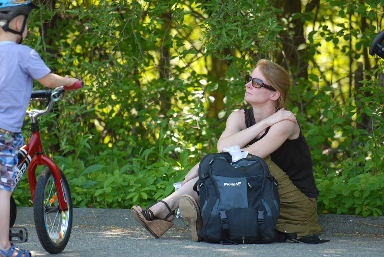 A woman sits on the ground with a backpack, wearing sunglasses, while a child on a bike stands nearby.