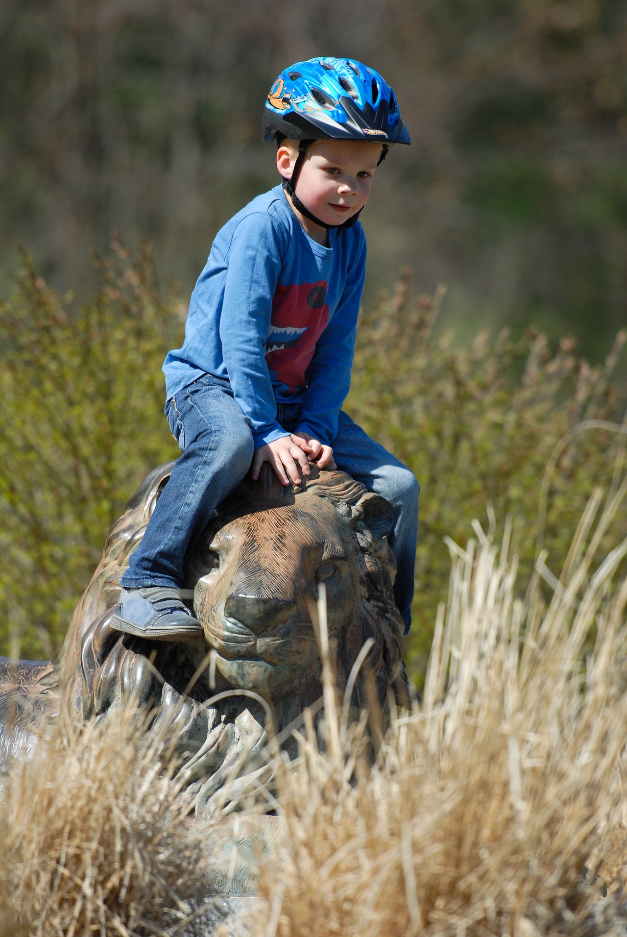 A child wearing a helmet sits on a lion statue outdoors, surrounded by tall grass and bushes.