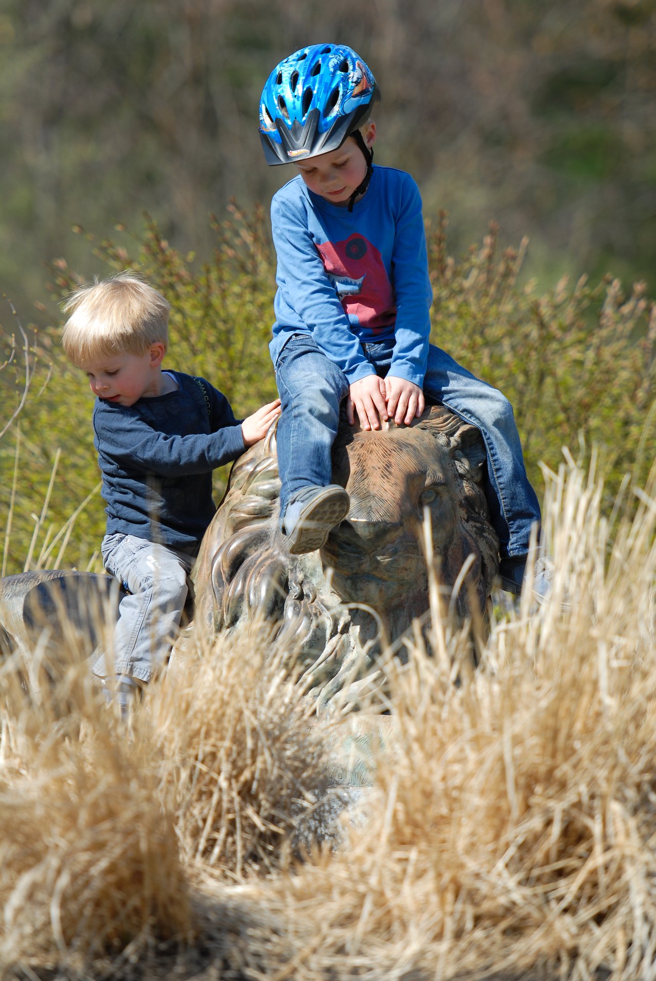 Two children climb and sit on a large lion statue outdoors.