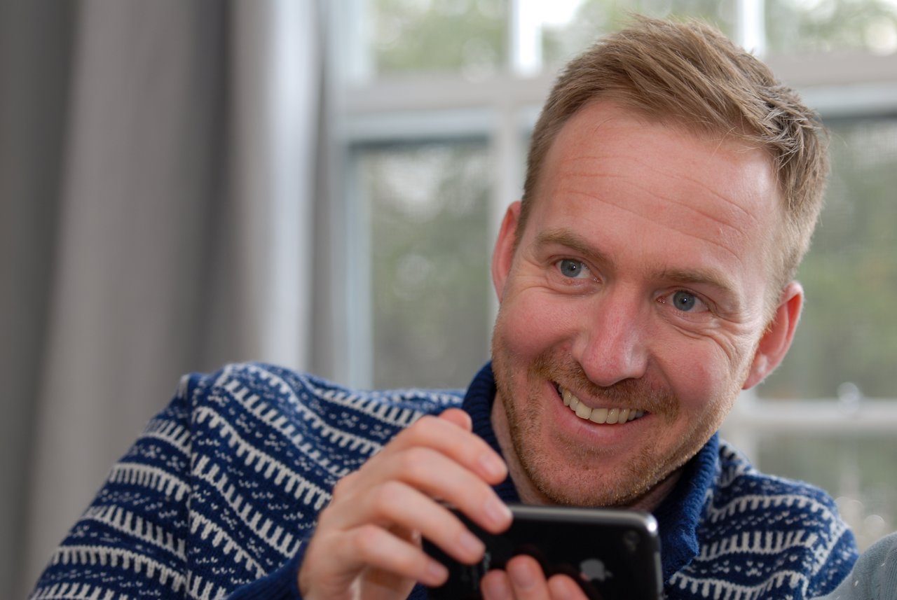 A smiling man in a blue sweater holds a smartphone and looks at something off-camera.