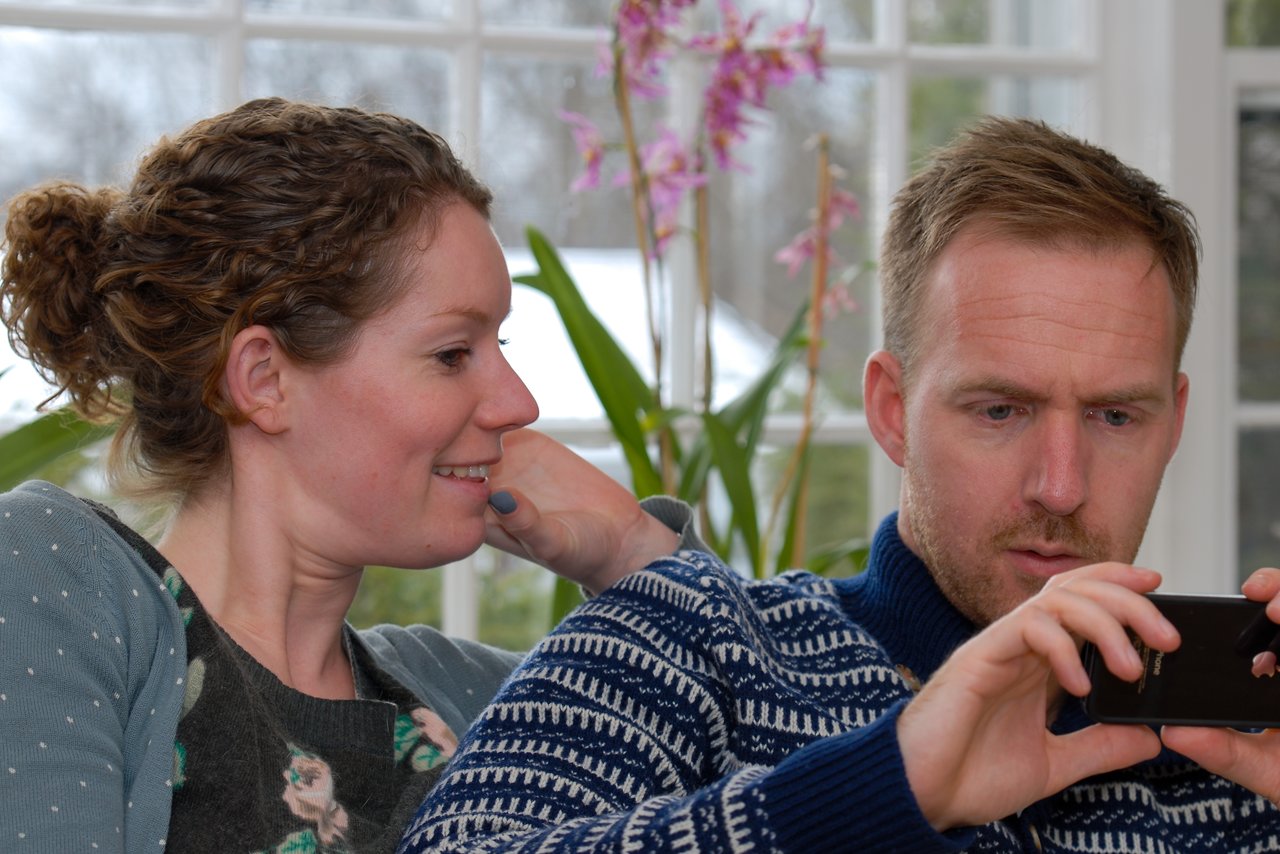 A woman smiles while watching a man focused on his phone, both sitting closely together indoors.