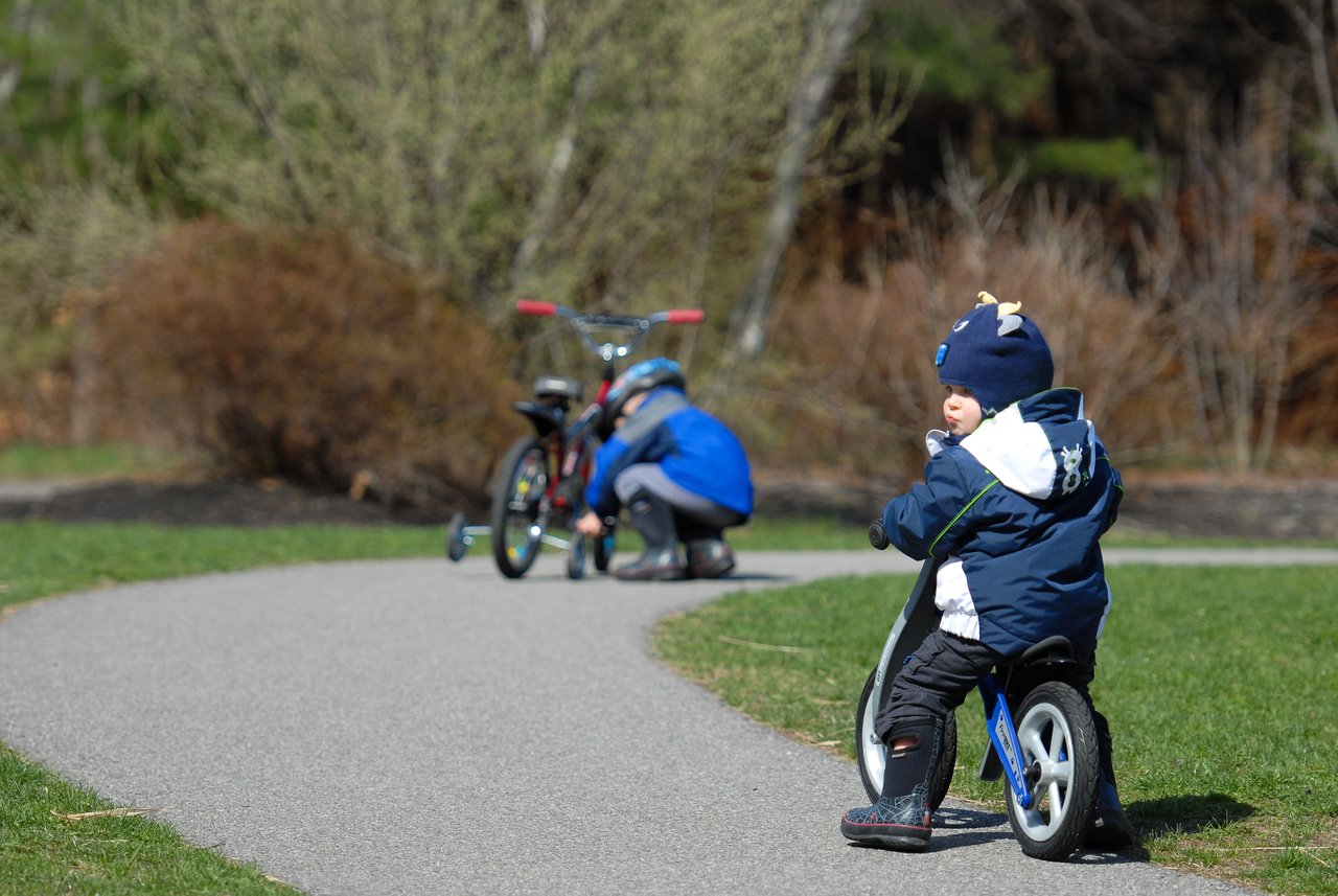 A child rides a small bike while another person kneels to adjust a bicycle on a paved path.