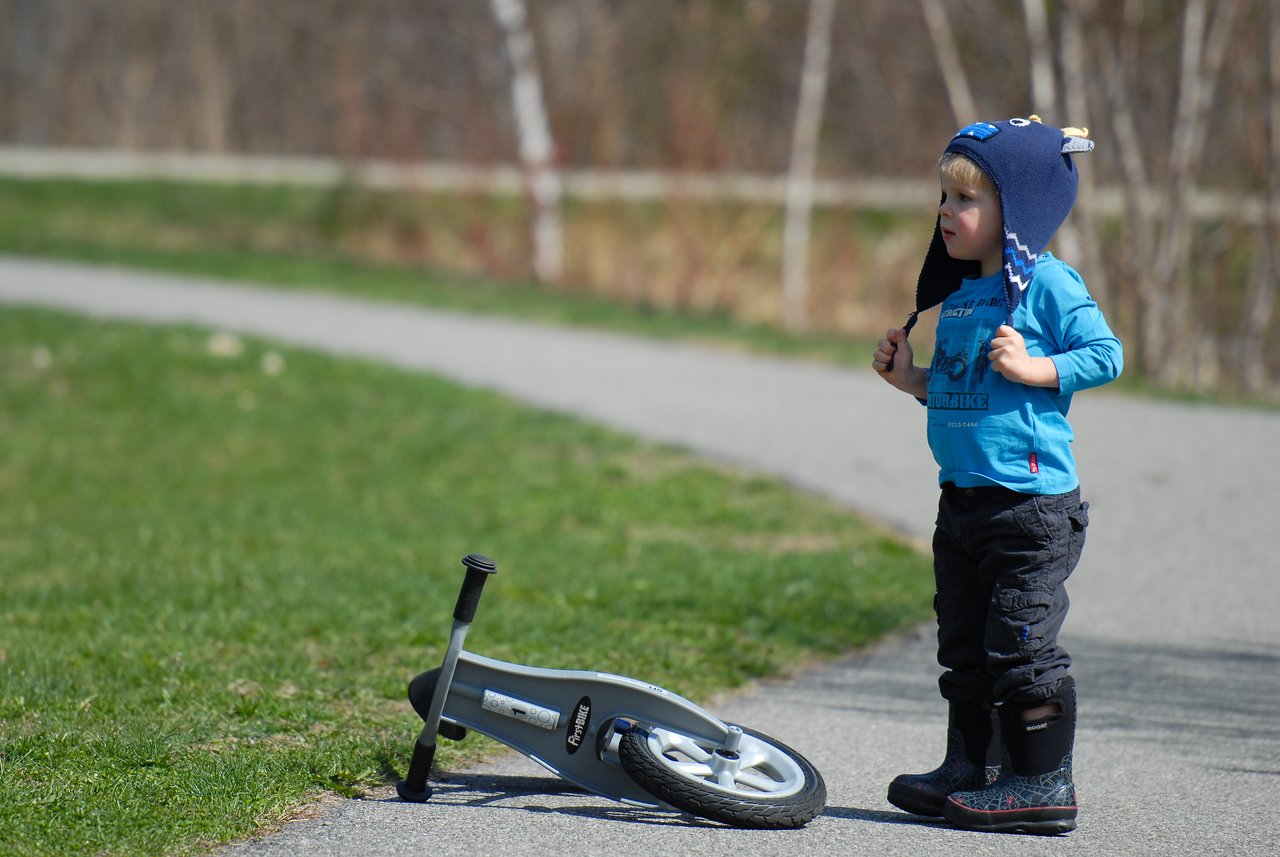 A young child stands on a paved path next to a fallen balance bike, looking ahead with a curious expression.