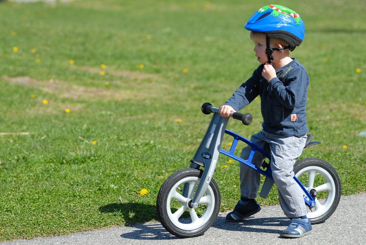A young child wearing a blue helmet rides a small balance bike on a paved path in a grassy area.