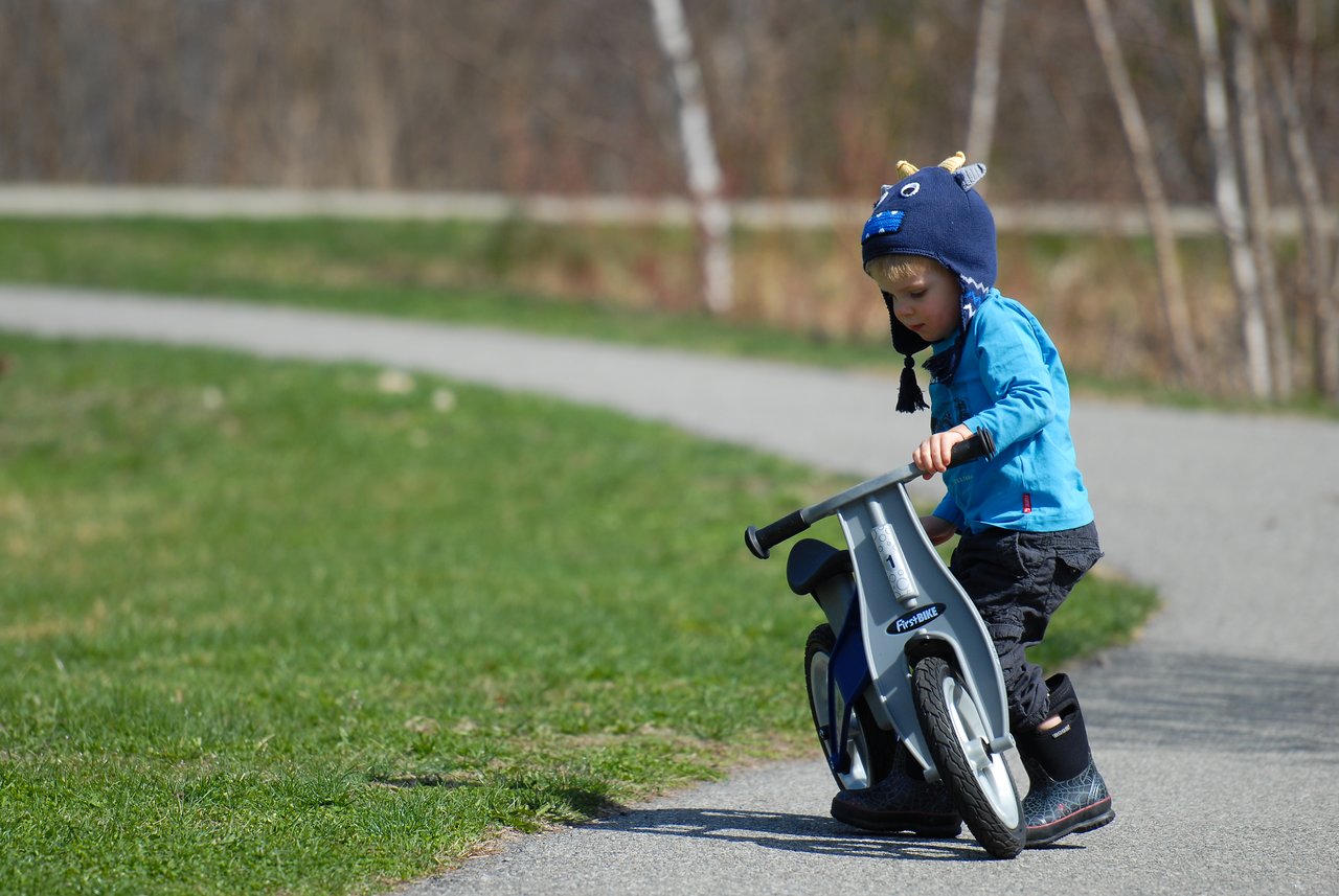 A young child wearing a blue jacket and hat rides a small balance bike on a paved path outdoors.