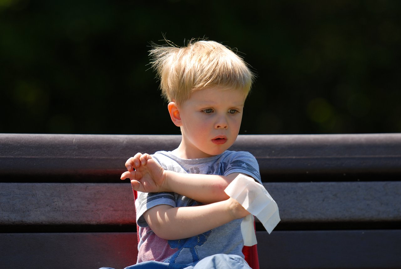 A young child sits on a bench, looking at their bandaged arm with a concerned expression.
