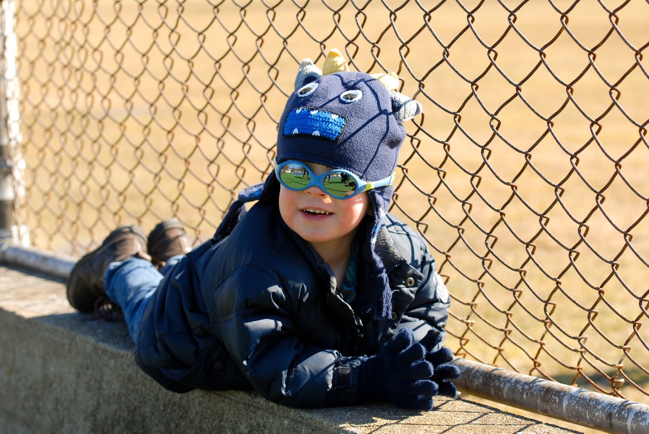 A child in a winter jacket and hat leans on a concrete ledge, smiling near a chain-link fence.