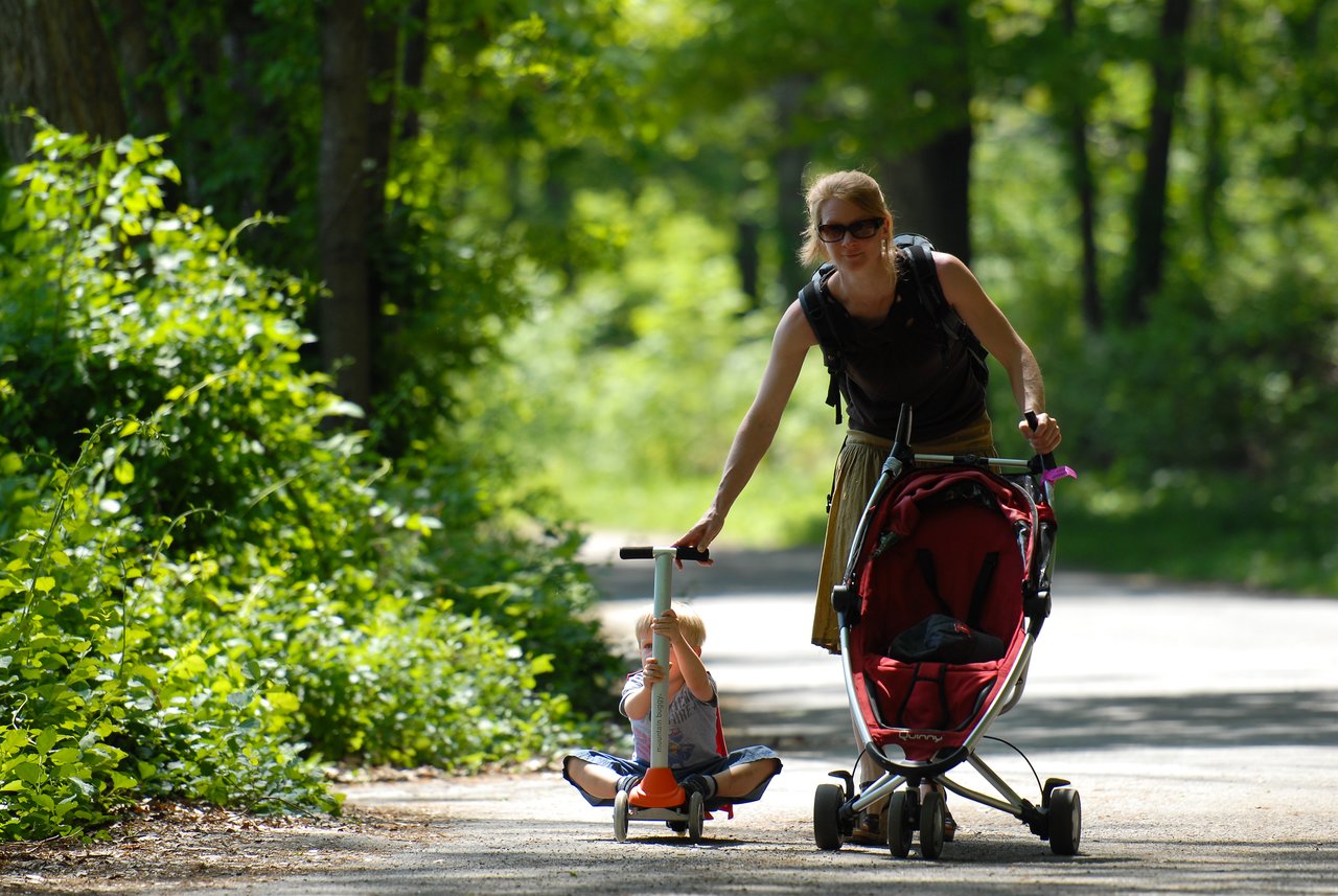 A young child rides a small scooter while an adult walks beside them, pushing an empty stroller.