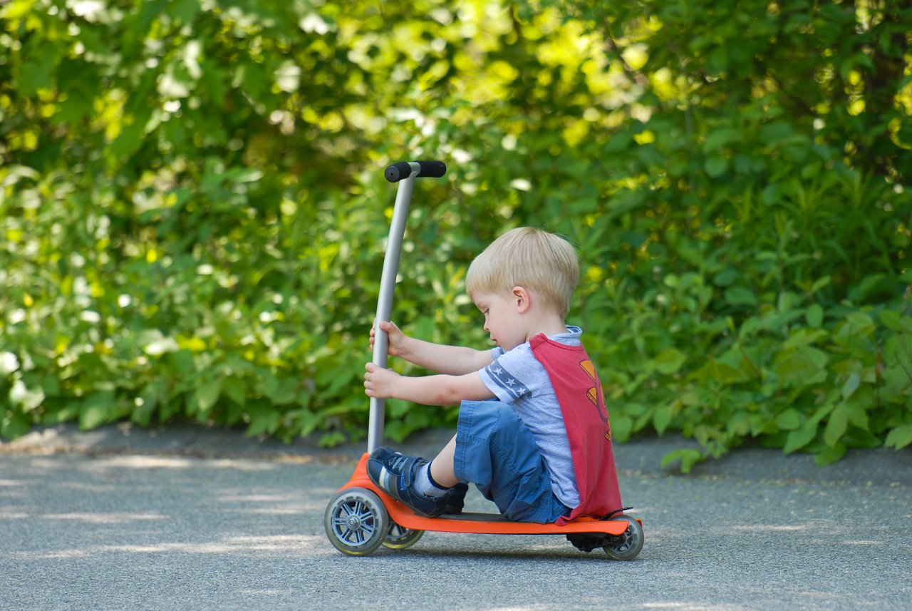 A young child sits on an orange scooter, holding the handlebars and pushing forward on a paved path.