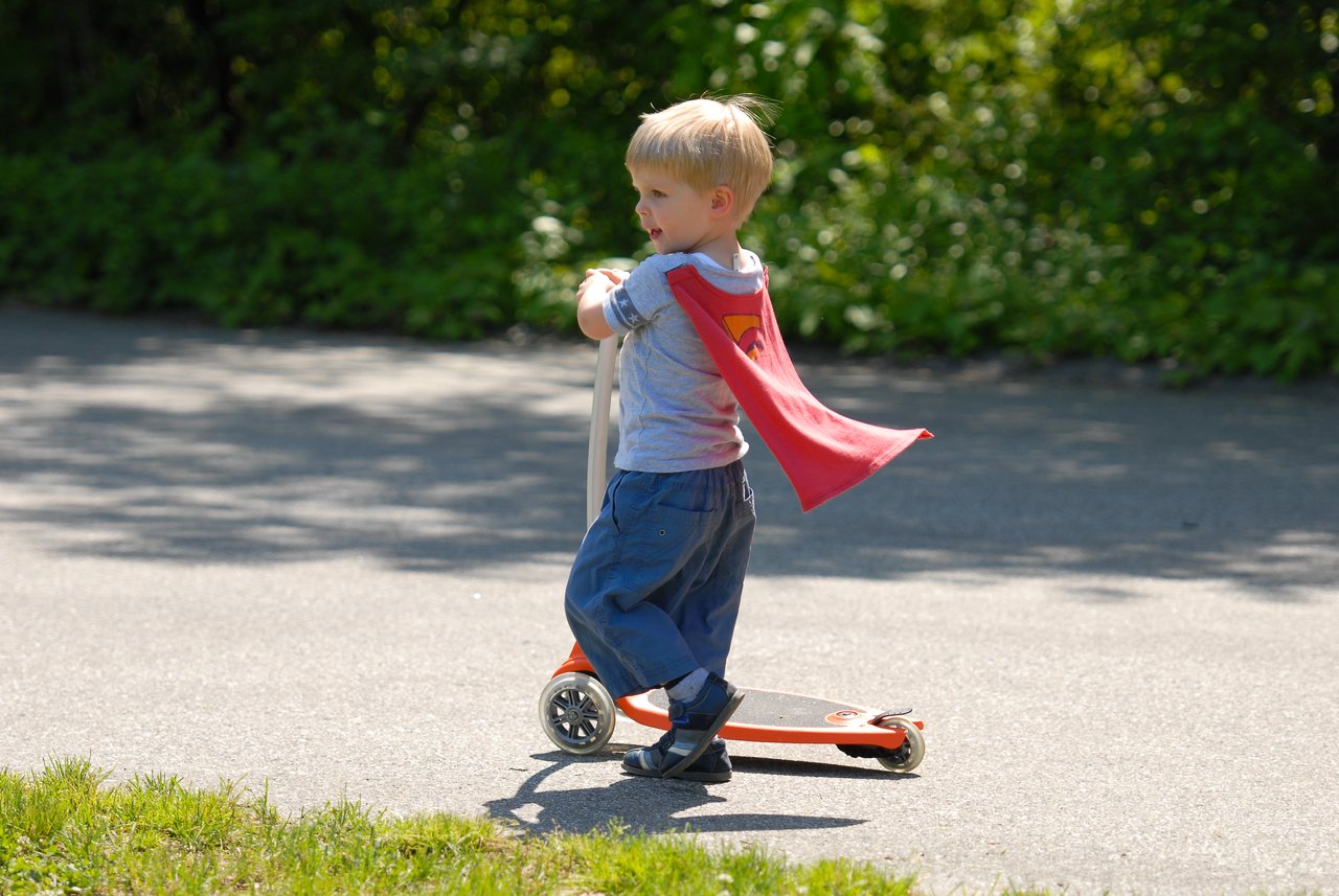 A young child wearing a red cape rides an orange scooter on a paved path outdoors.