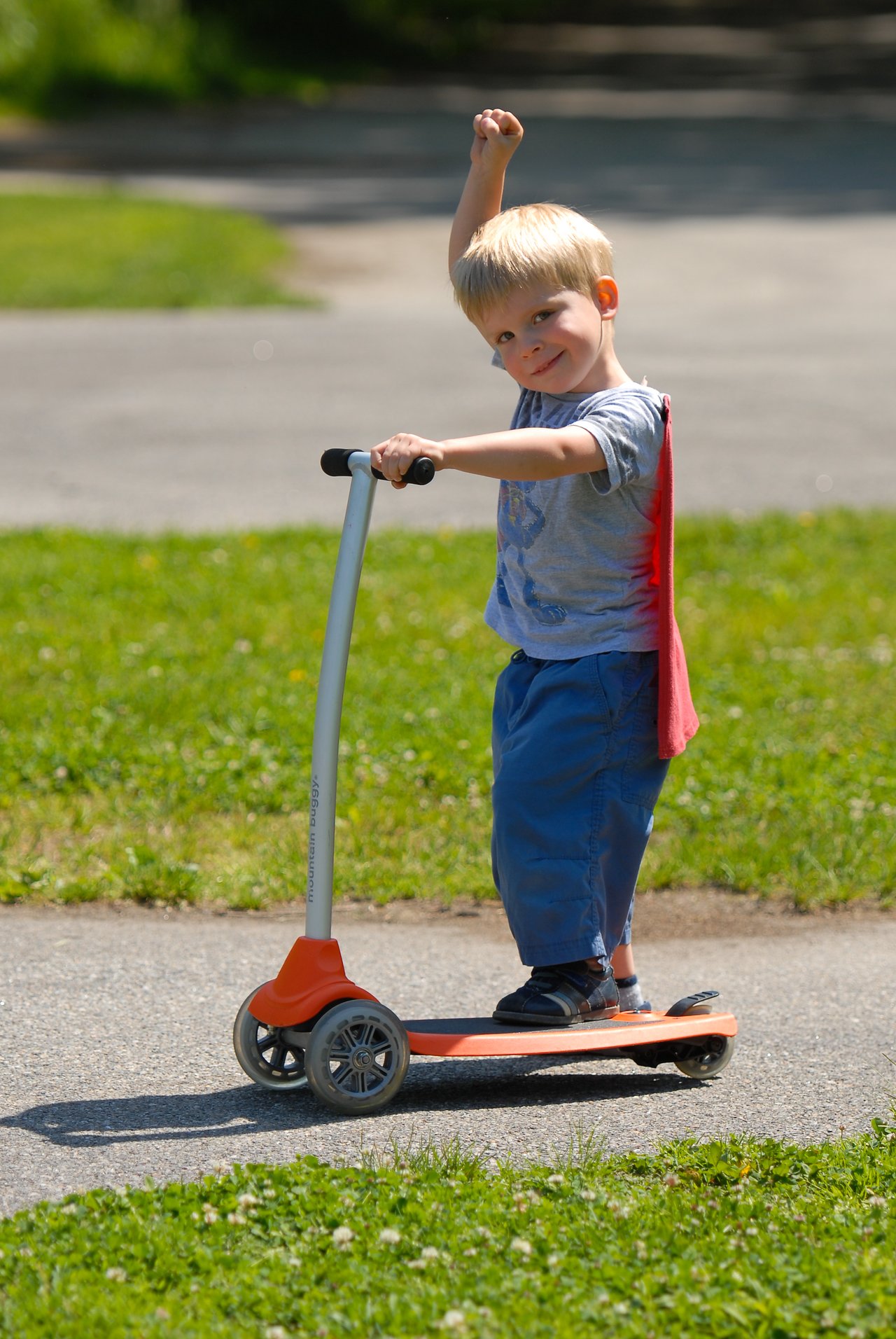 A young child rides an orange scooter on a paved path, wearing a red cape and raising one arm.