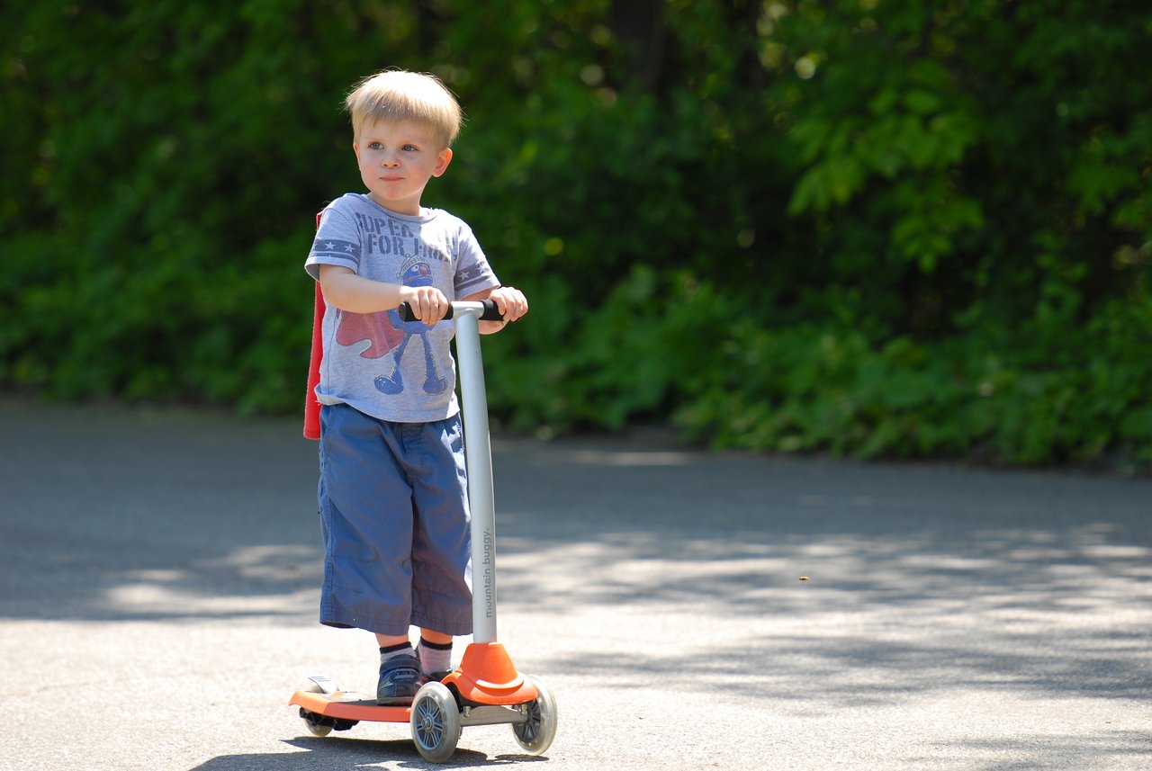 A young child rides a three-wheeled scooter on a paved path, wearing a t-shirt, shorts, and a cape.