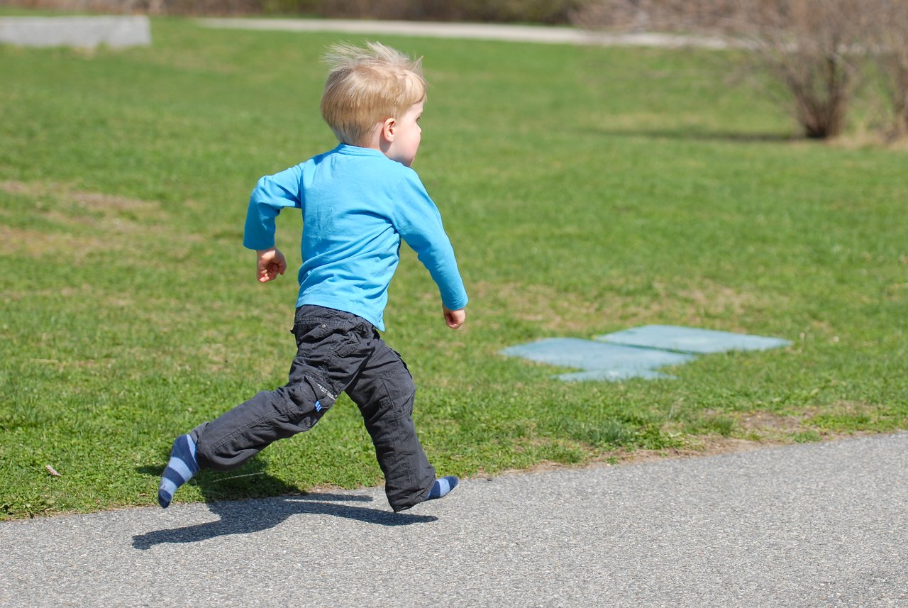 A young child in a blue shirt and dark pants runs on a paved path in a grassy area.