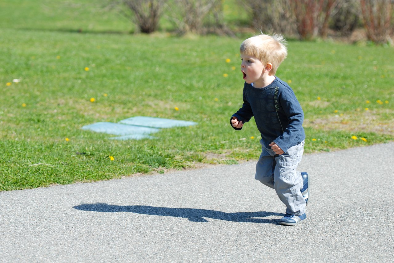 A young child with blond hair runs on a paved path, wearing a blue shirt and light-colored pants.