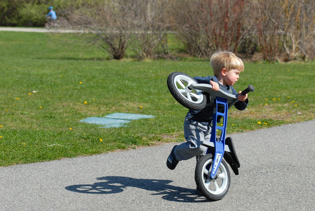 A young child lifts and tilts a small balance bike while walking on a paved path in a park.