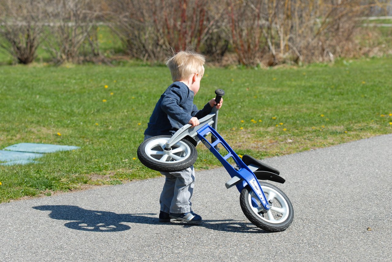 A young child lifts a small blue balance bike off the ground while standing on a paved path.